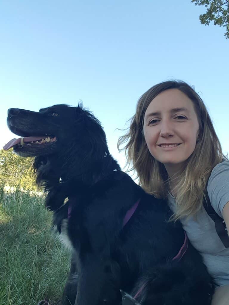 Selfie joyeux : femme et chien noir souriants sous le ciel Femme souriante et chien noir avec harnais violet, langue pendante, posant en selfie sous un ciel bleu clair.