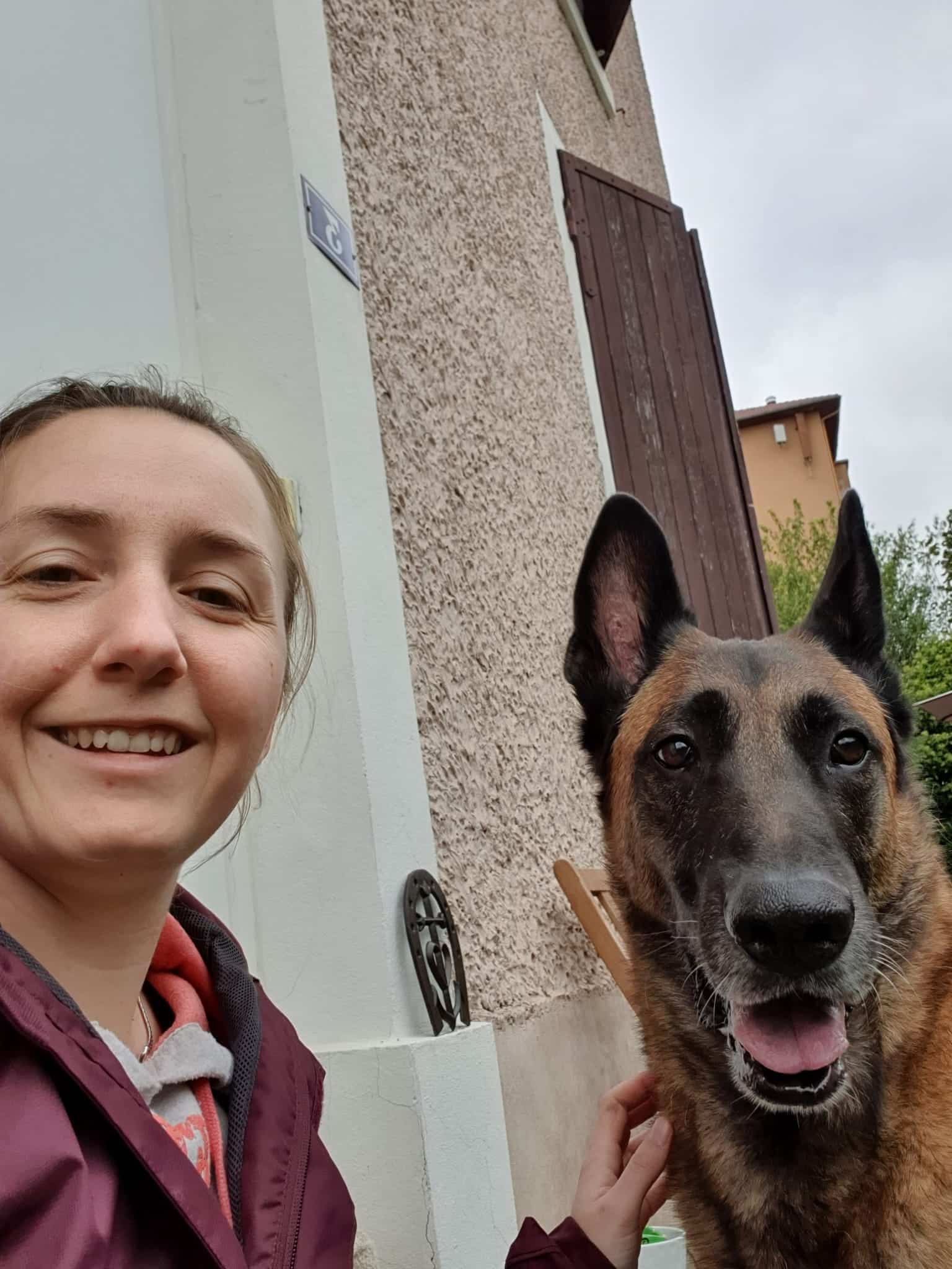 Femme souriante et Malinois joyeux posant devant une façade de maison texturée, le numéro 3 visible sur une plaque.