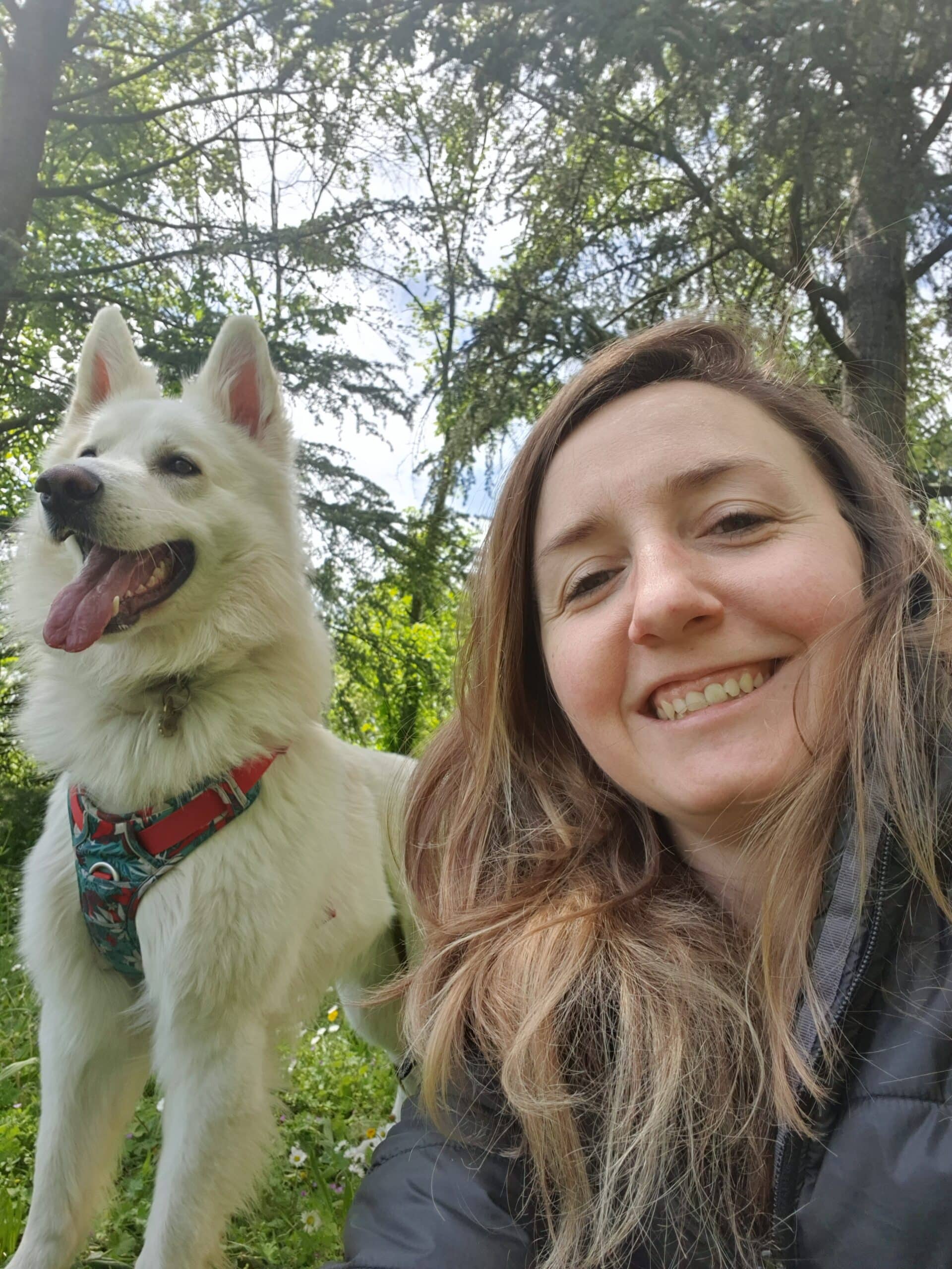 Femme souriante aux cheveux bruns avec son grand chien blanc à la langue pendante, posant en selfie dans une forêt verdoyante.