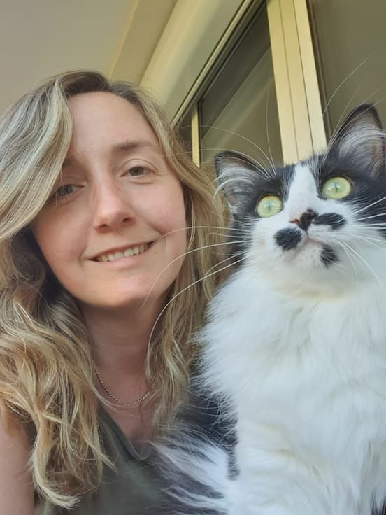 Selfie Complice : Femme Souriante et Chat Noir & Blanc Unique Femme blonde souriante et son chat noir et blanc aux yeux verts brillants et moustache distinctive en selfie.